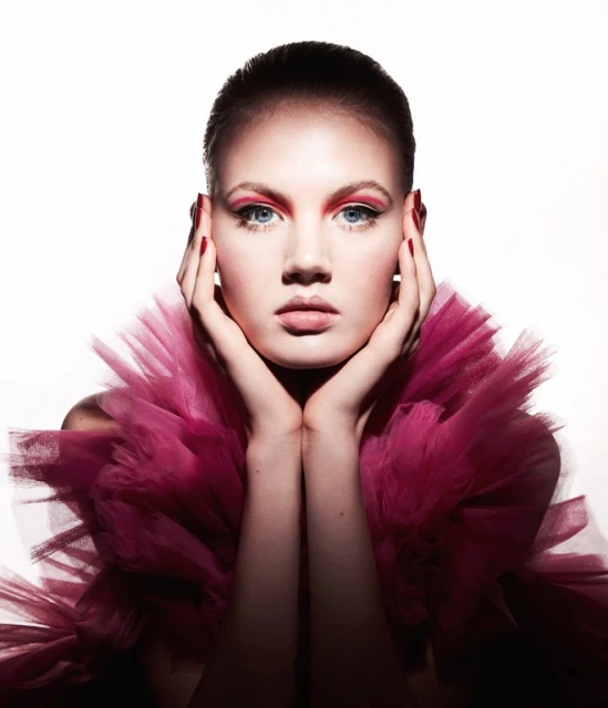 Close-up portrait of a young adult female model with pink eye makeup, full lips, and a pink tulle ruffled collar on a white background.