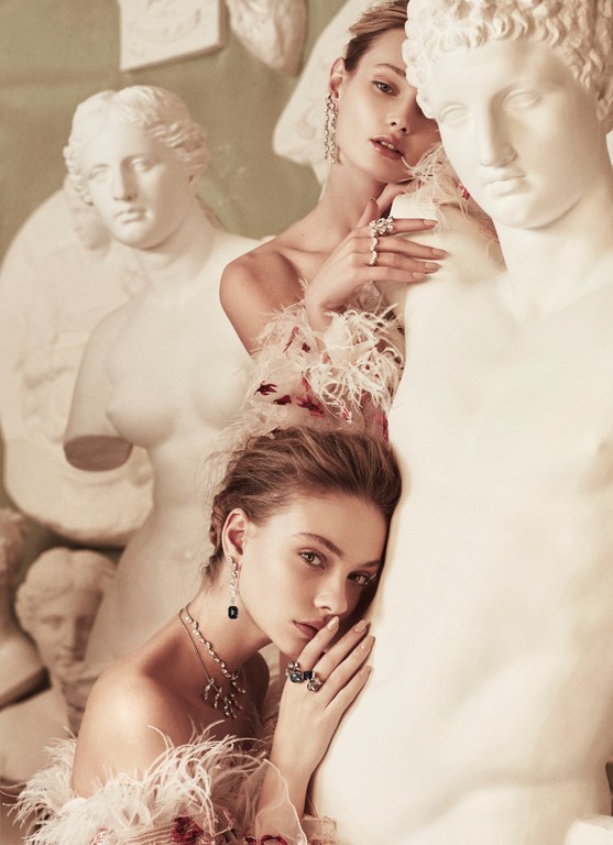 Two women in feathered off-the-shoulder tops pose with classical statues in a soft, vintage-toned studio setting for a fashion editorial.