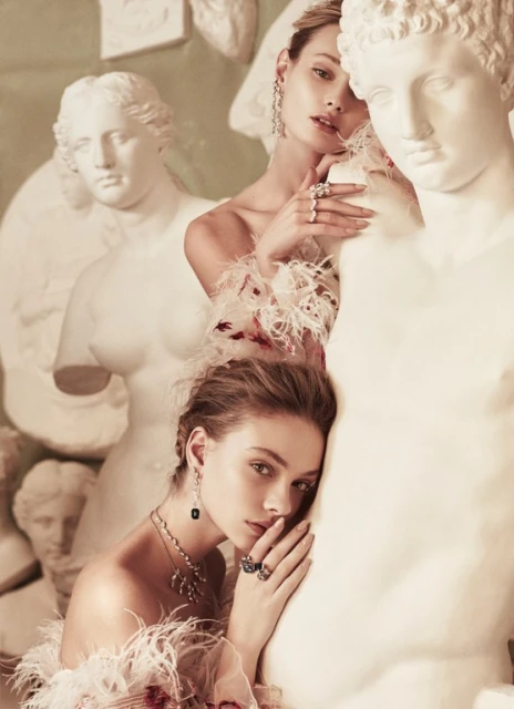 Two women in feathered off-the-shoulder tops pose with classical statues in a soft, vintage-toned studio setting for a fashion editorial.