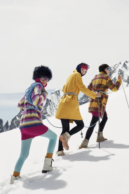 Three women in colorful winter coats and boots hike up a snowy mountain with a mountain range in the background on a bright day.