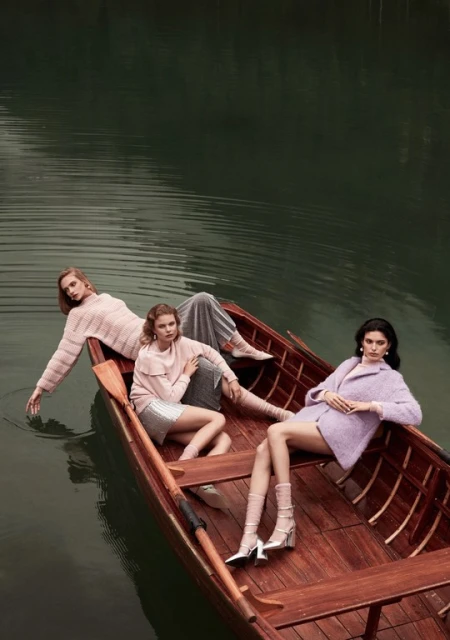 Three fashion models lounge in a wooden rowboat on a lake surrounded by trees, wearing pastel colored knitwear and dresses.