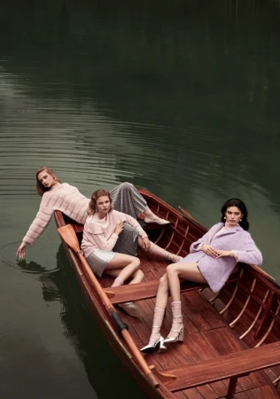 Three fashion models lounge in a wooden rowboat on a lake surrounded by trees, wearing pastel colored knitwear and dresses.