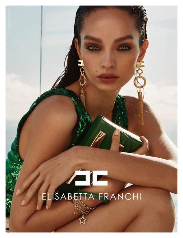 Close-up of a woman with long brown hair, green eyes, wearing a green sequin top and gold earrings, holding a green purse, in a studio.