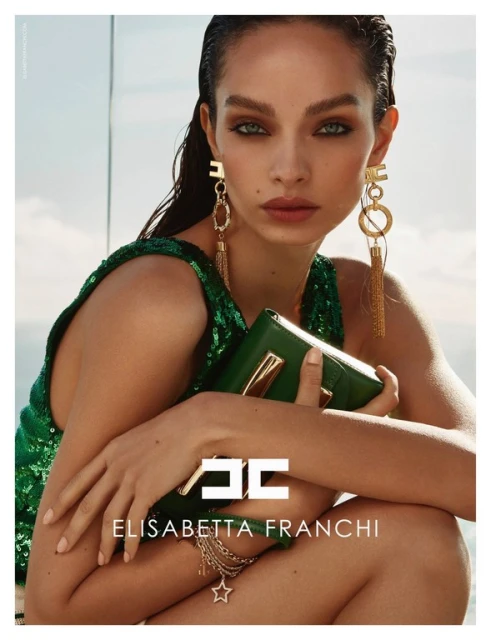 Close-up of a woman with long brown hair, green eyes, wearing a green sequin top and gold earrings, holding a green purse, in a studio.
