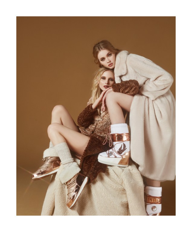 Two women in fur coats and winter boots pose against a neutral backdrop. Fashion shot featuring matching textures and warm, tonal colors.