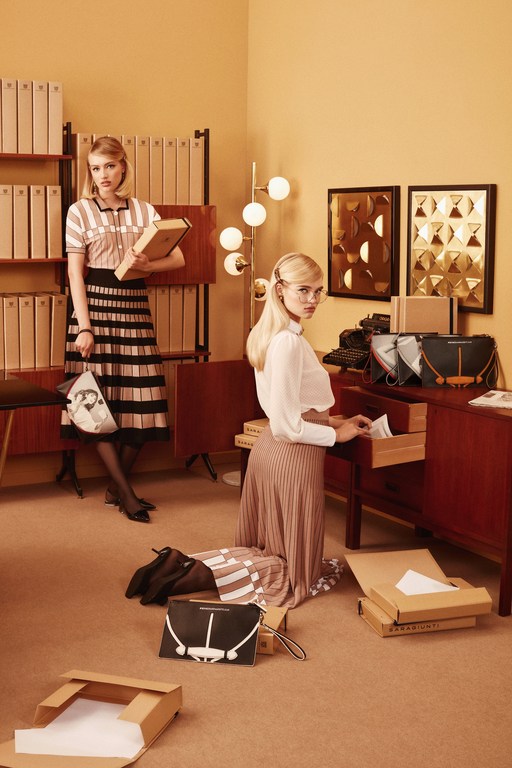 Two women in a retro office organize handbags and paperwork. One woman stands with a book and bag; the other kneels by a desk.