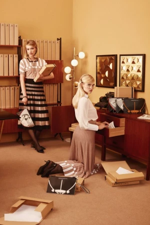 Two women in a retro office organize handbags and paperwork. One woman stands with a book and bag; the other kneels by a desk.