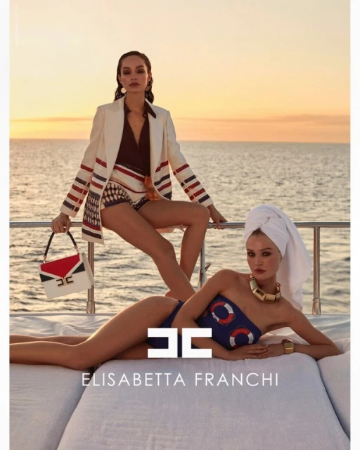 Two women pose in swimwear and a blazer on a yacht against a sunset backdrop, showcasing fashion and summer lifestyle.