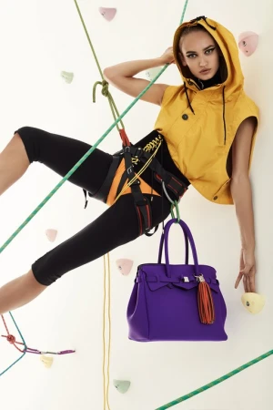Woman in a yellow hooded vest poses on a climbing wall with a purple handbag hanging from her harness in a fashion editorial shot.