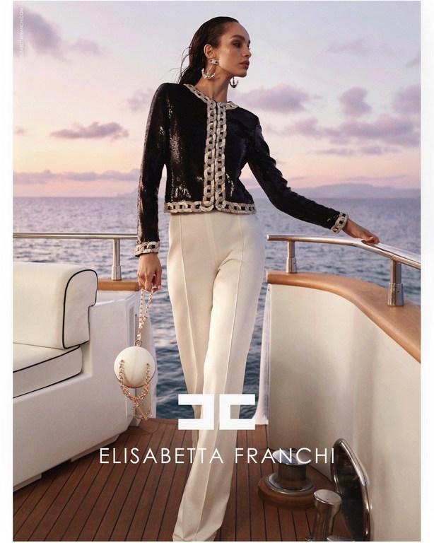 Elegant woman poses on yacht in Elisabetta Franchi fashion. Black sequin jacket, white pants, round handbag, ocean background at sunset.