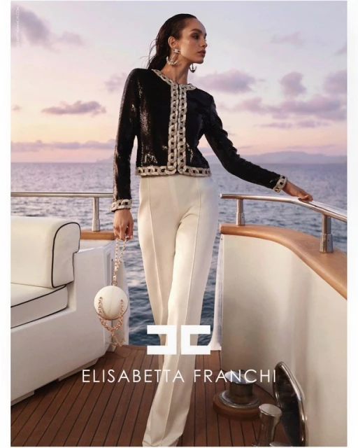 Elegant woman poses on yacht in Elisabetta Franchi fashion. Black sequin jacket, white pants, round handbag, ocean background at sunset.