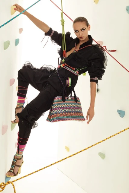 Woman in black jumpsuit and high heels scaling a climbing wall indoors, holding a patterned purse, with climbing ropes around her.