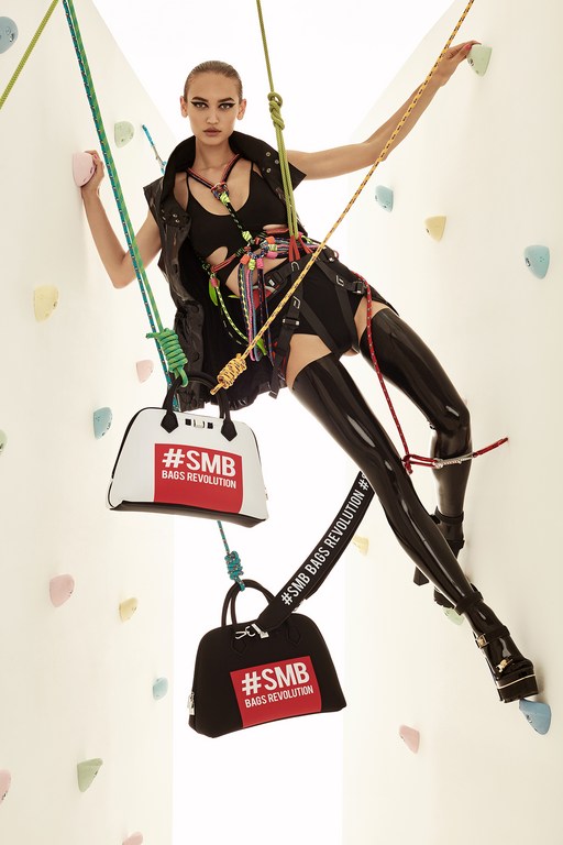A fashion shot of a young adult woman rock climbing indoors, modeling in a black outfit and platform shoes, with bags attached to the harness.