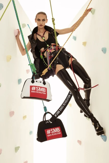 A fashion shot of a young adult woman rock climbing indoors, modeling in a black outfit and platform shoes, with bags attached to the harness.