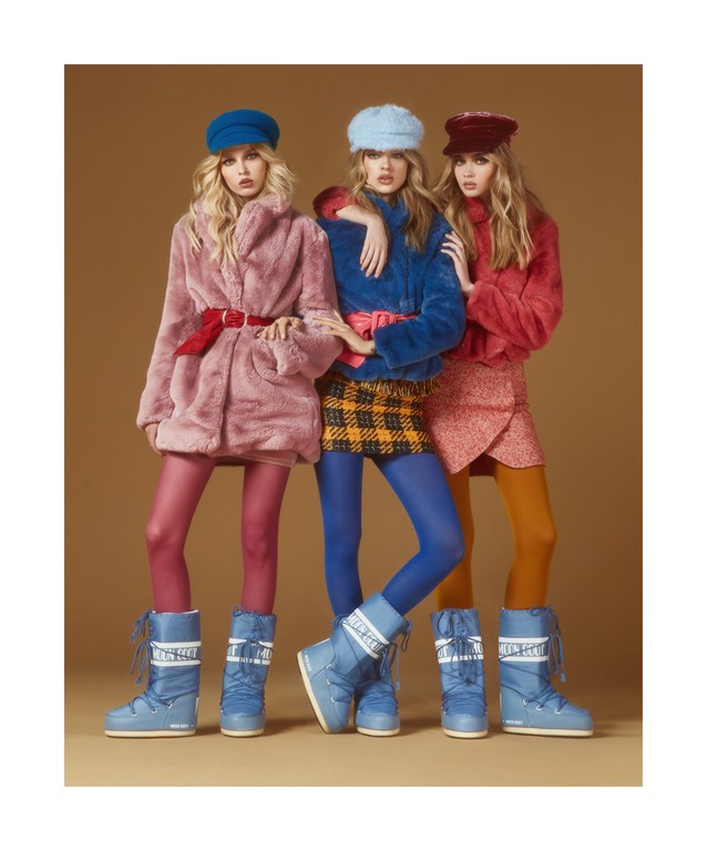 Three fashion models pose in colorful winter outfits: furry coats, hats, tights, and moon boots, against a tan studio backdrop.