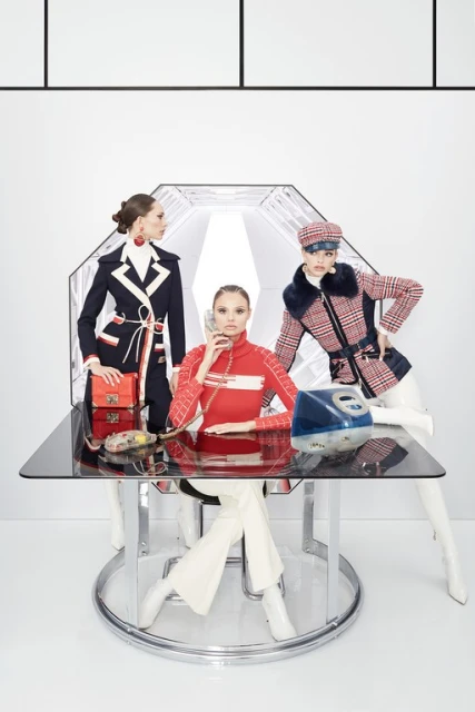 Three fashion models pose at a glass table, one talking on a retro phone in a white interior, wearing 1960s-inspired outfits.
