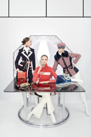 Three fashion models pose at a glass table, one talking on a retro phone in a white interior, wearing 1960s-inspired outfits.