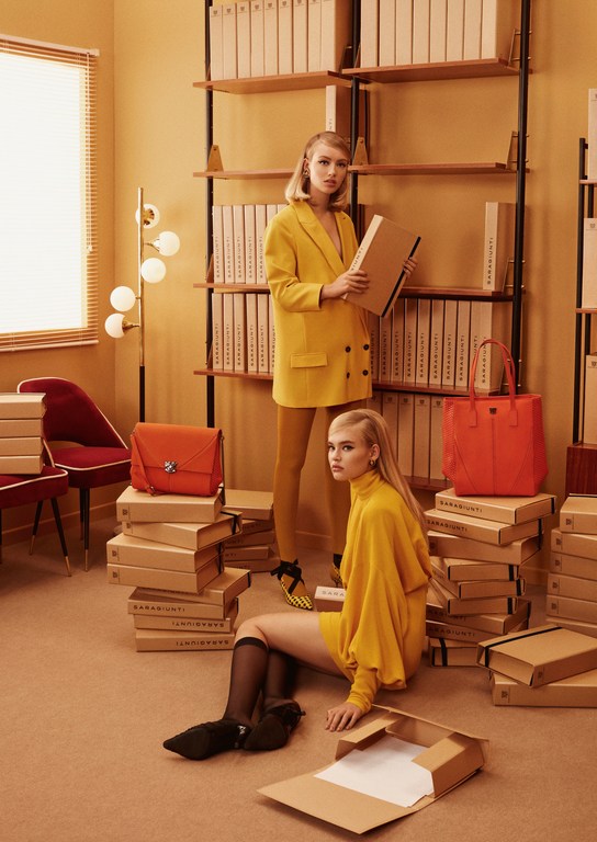 Two blond models in yellow clothing pose in a room with a bookshelf and stacks of boxes, creating a retro fashion editorial.