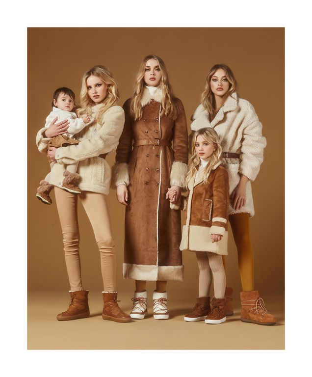 A group of four blonde women, a baby and a girl are standing together, wearing matching vintage-style coats and winter boots in studio lighting.
