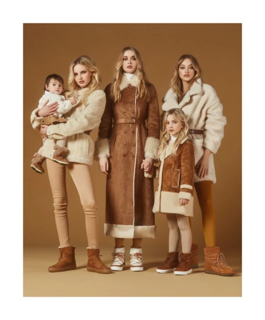 A group of four blonde women, a baby and a girl are standing together, wearing matching vintage-style coats and winter boots in studio lighting.