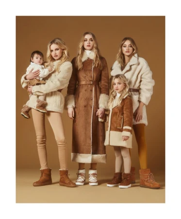 A group of four blonde women, a baby and a girl are standing together, wearing matching vintage-style coats and winter boots in studio lighting.