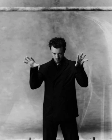 Monochrome portrait of a man with curly hair in a dark suit against a textured gray backdrop with light flares.