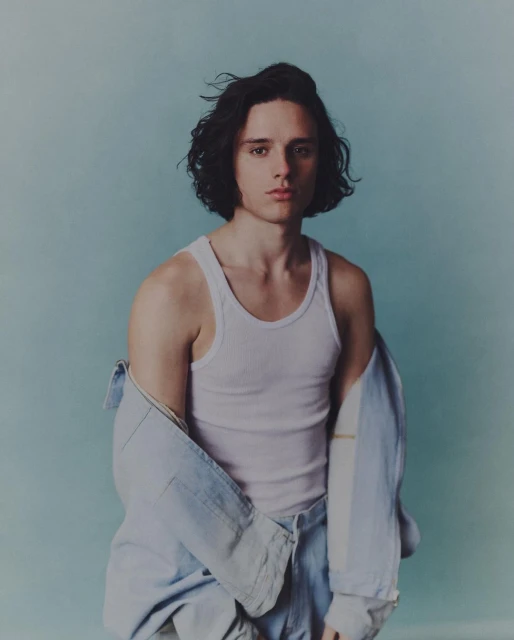 Portrait of a young adult man with dark curly hair wearing a white tank top and an unbuttoned light blue denim shirt against a light blue background.