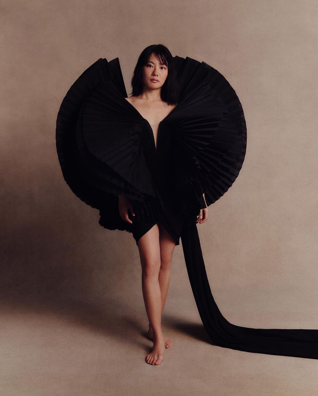 Fashionable woman poses barefoot in an avant-garde black dress with full, pleated detailing in a studio setting, spotlighted in a fashion shot.