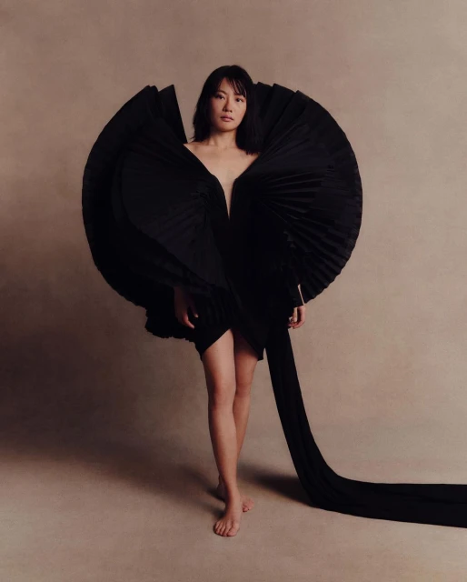 Fashionable woman poses barefoot in an avant-garde black dress with full, pleated detailing in a studio setting, spotlighted in a fashion shot.