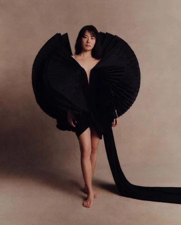 Fashionable woman poses barefoot in an avant-garde black dress with full, pleated detailing in a studio setting, spotlighted in a fashion shot.