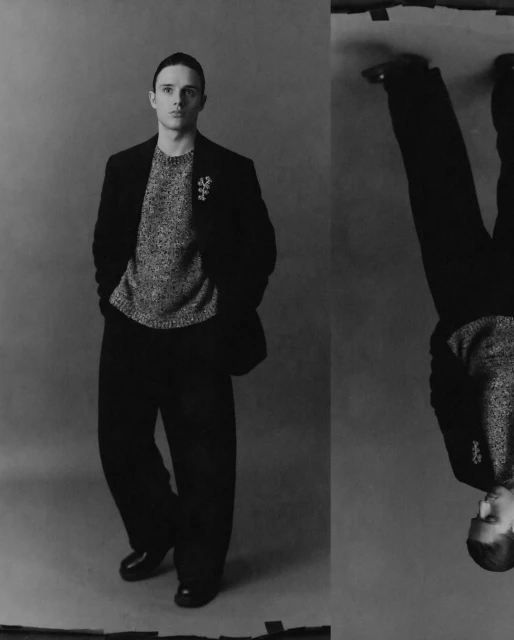 Black and white portrait of a young adult male in a sweater, blazer, and wide-leg trousers, presented with a mirrored, upside-down duplicate.