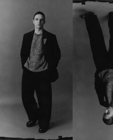 Black and white portrait of a young adult male in a sweater, blazer, and wide-leg trousers, presented with a mirrored, upside-down duplicate.
