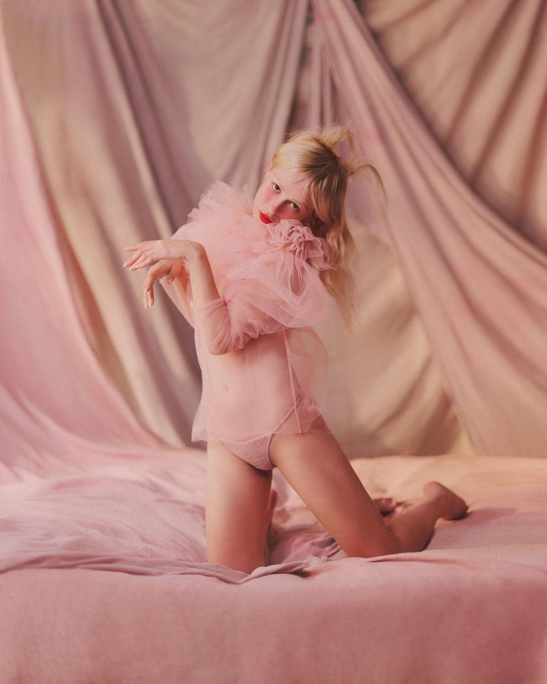 Young woman kneeling on a pink bed wearing a sheer pink outfit with a large tulle ruffle collar, and a pink draped background.