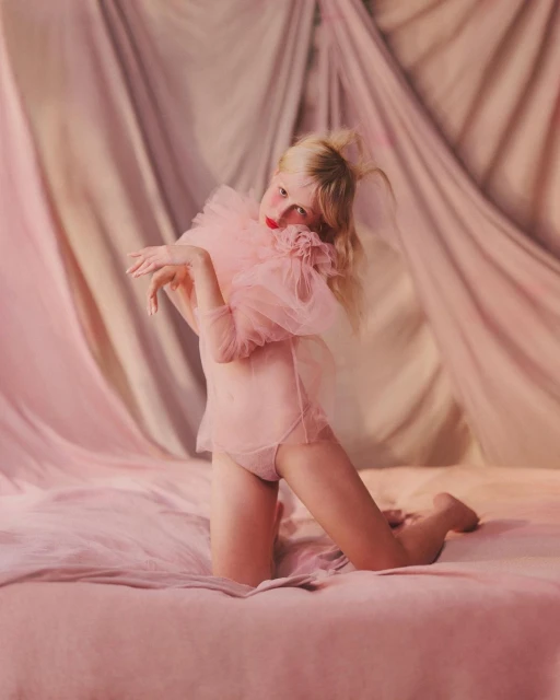 Young woman kneeling on a pink bed wearing a sheer pink outfit with a large tulle ruffle collar, and a pink draped background.