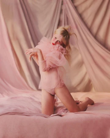 Young woman kneeling on a pink bed wearing a sheer pink outfit with a large tulle ruffle collar, and a pink draped background.