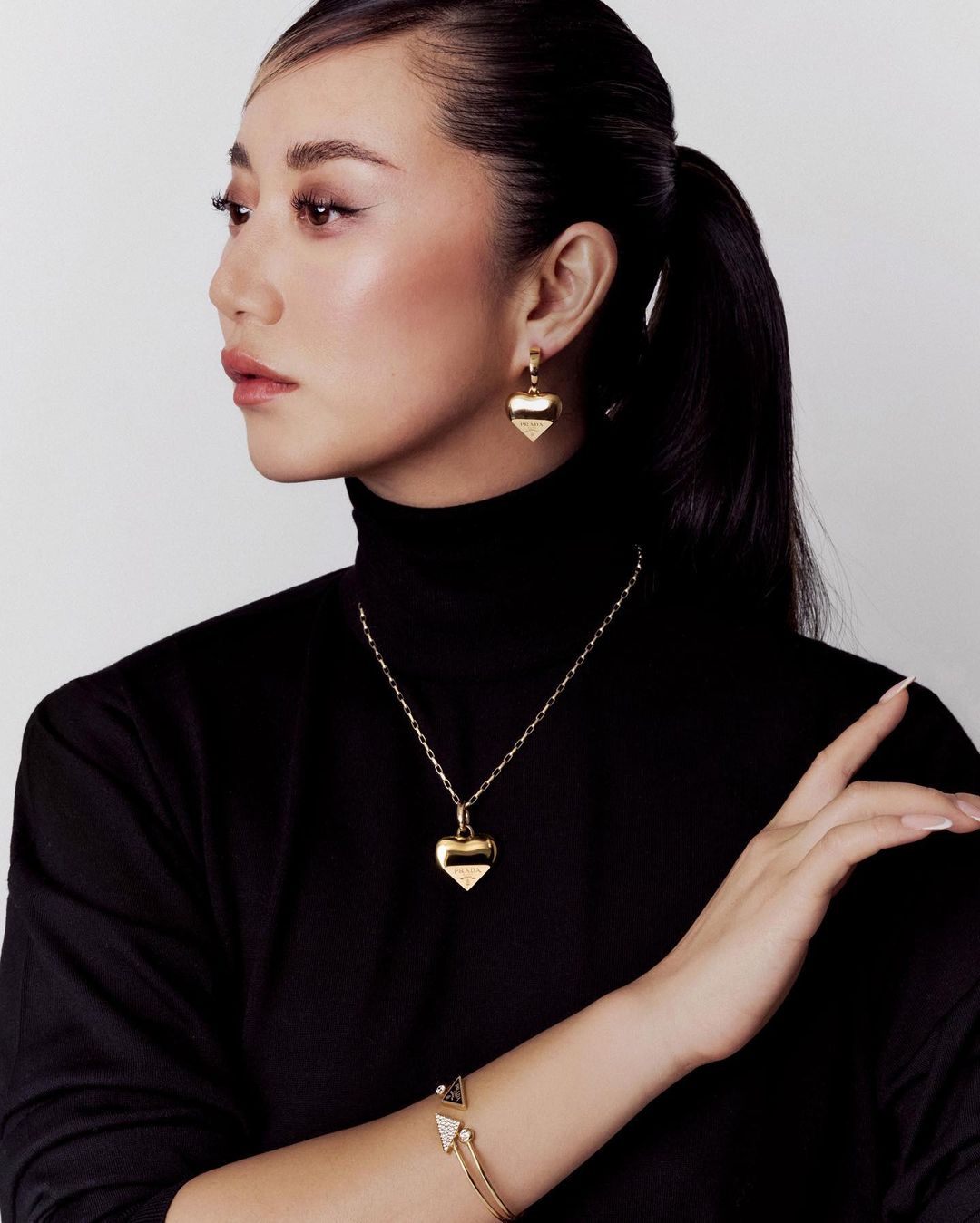 Glamour shot of a woman with her dark hair in a ponytail, wearing a black turtleneck, gold heart earrings, necklace and a bracelet.