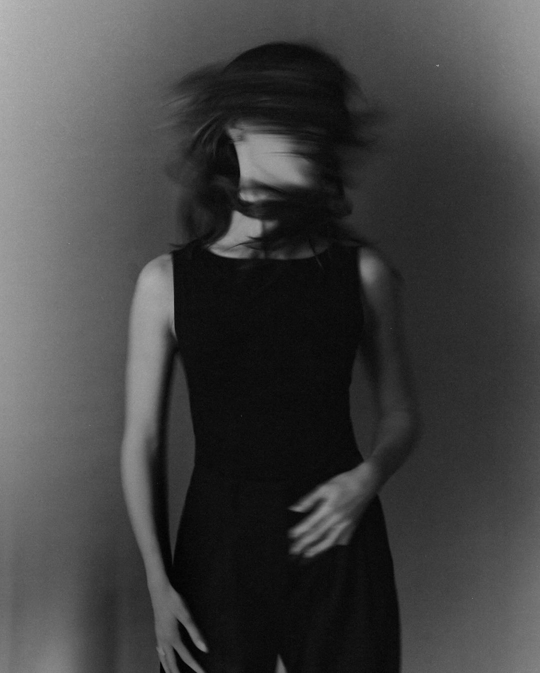 Black-and-white portrait of a woman in a black dress with blurred motion in her hair, set against a neutral background.