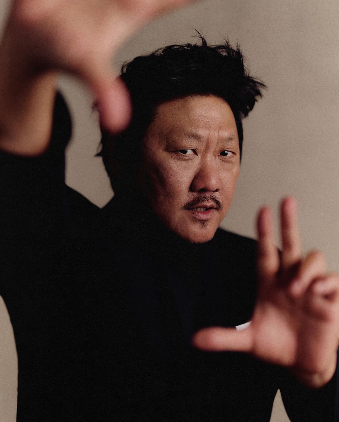 Headshot of Asian adult male in black turtleneck making framing gesture against plain backdrop in portrait orientation.