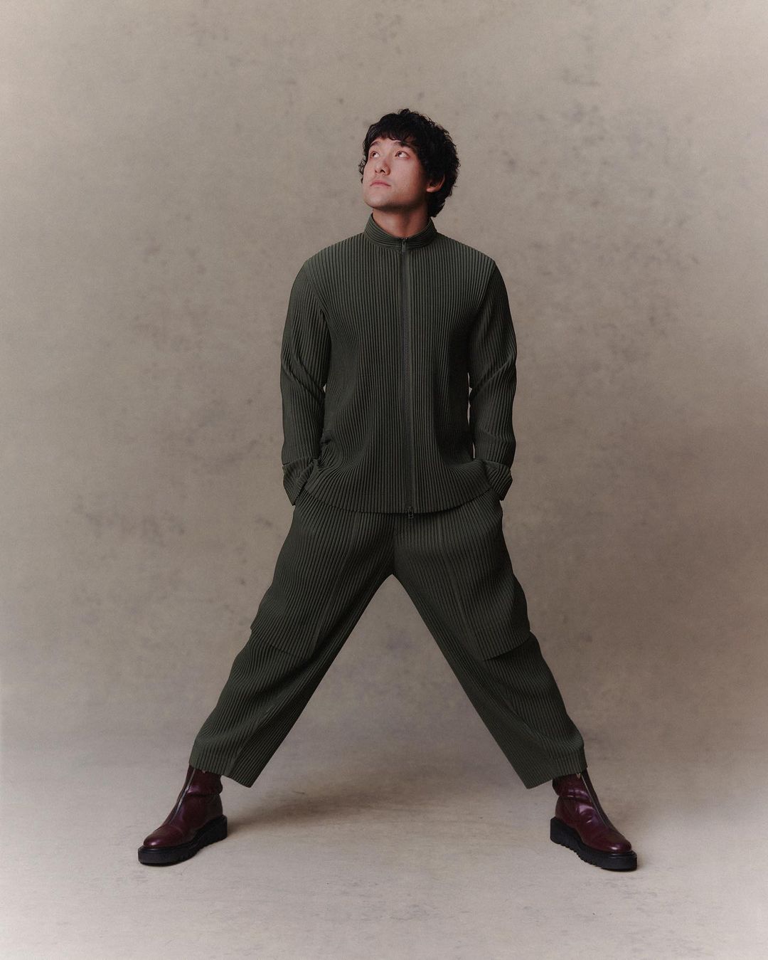 Portrait of an adult man in pleated matching set with brown ankle boots looking upwards in a studio setting with a neutral background.