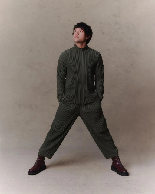 Portrait of an adult man in pleated matching set with brown ankle boots looking upwards in a studio setting with a neutral background.