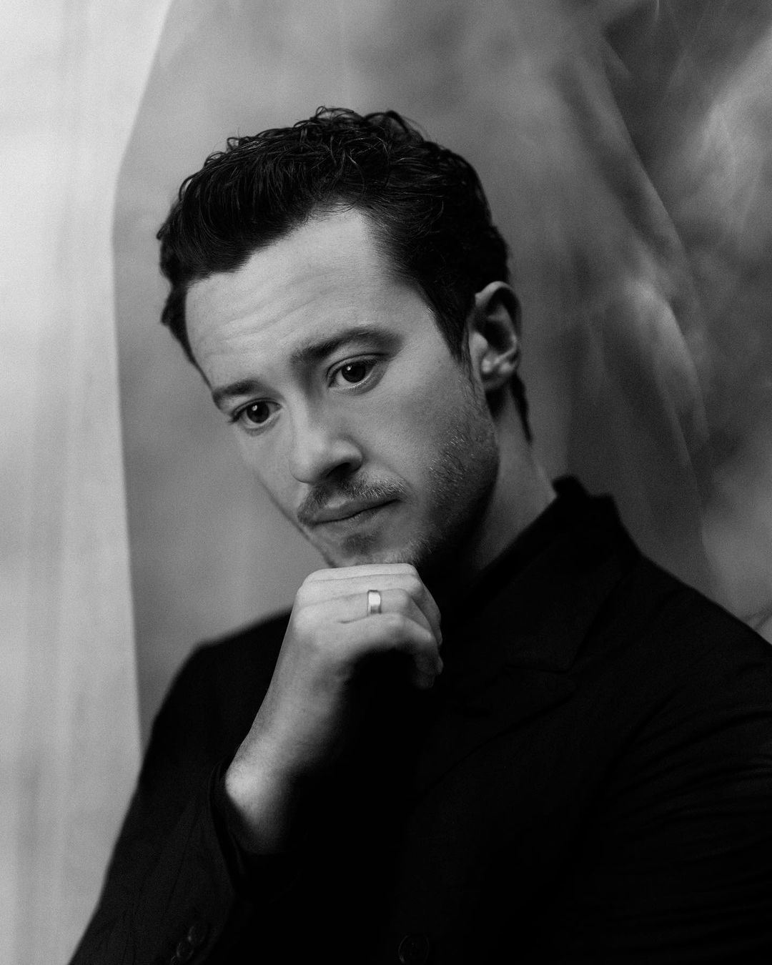 Portrait of an adult male with dark hair and a slight beard, wearing a dark suit, posing with his hand on his chin against a blurred background.