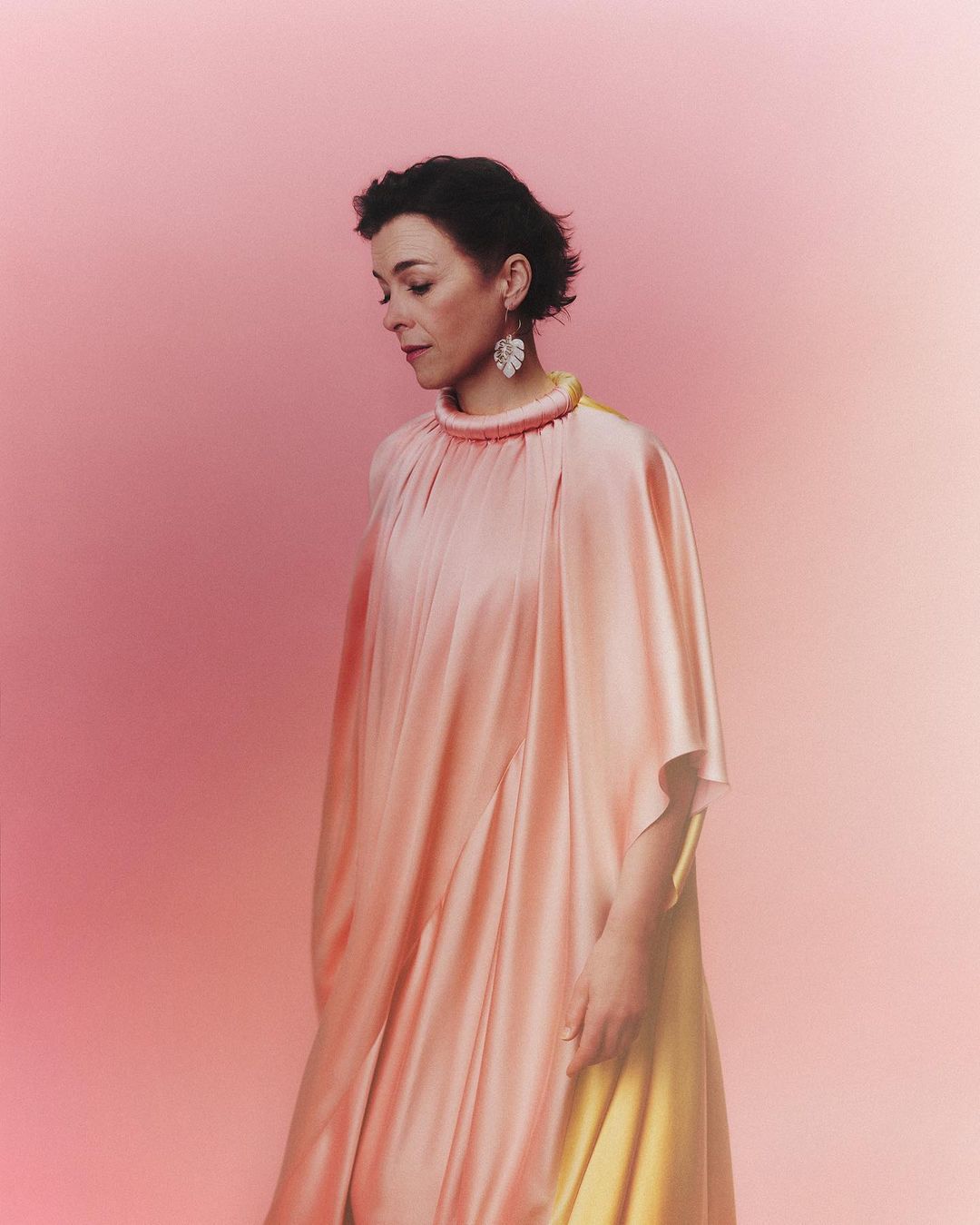 Portrait of a woman with short brown hair wearing a peach-colored dress and large earrings against a gradient pink background in a studio.