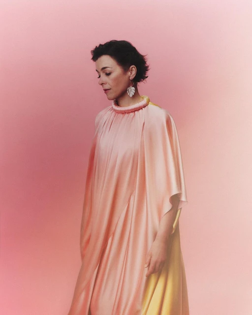 Portrait of a woman with short brown hair wearing a peach-colored dress and large earrings against a gradient pink background in a studio.