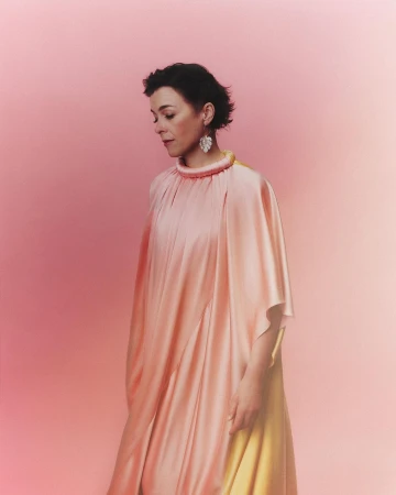 Portrait of a woman with short brown hair wearing a peach-colored dress and large earrings against a gradient pink background in a studio.