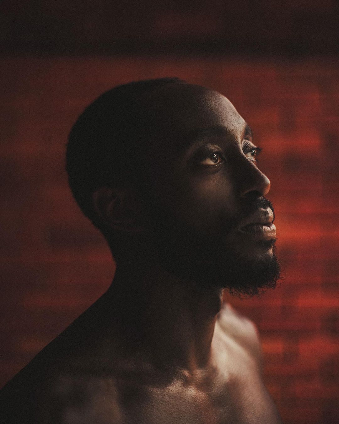 Portrait of a young, dark-skinned adult male with a beard and short hair looking upward against a textured reddish-brown background.