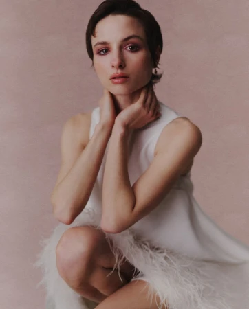 Close-up portrait of a woman with short dark hair wearing a white sleeveless dress with feather hem detail against a textured pink background.