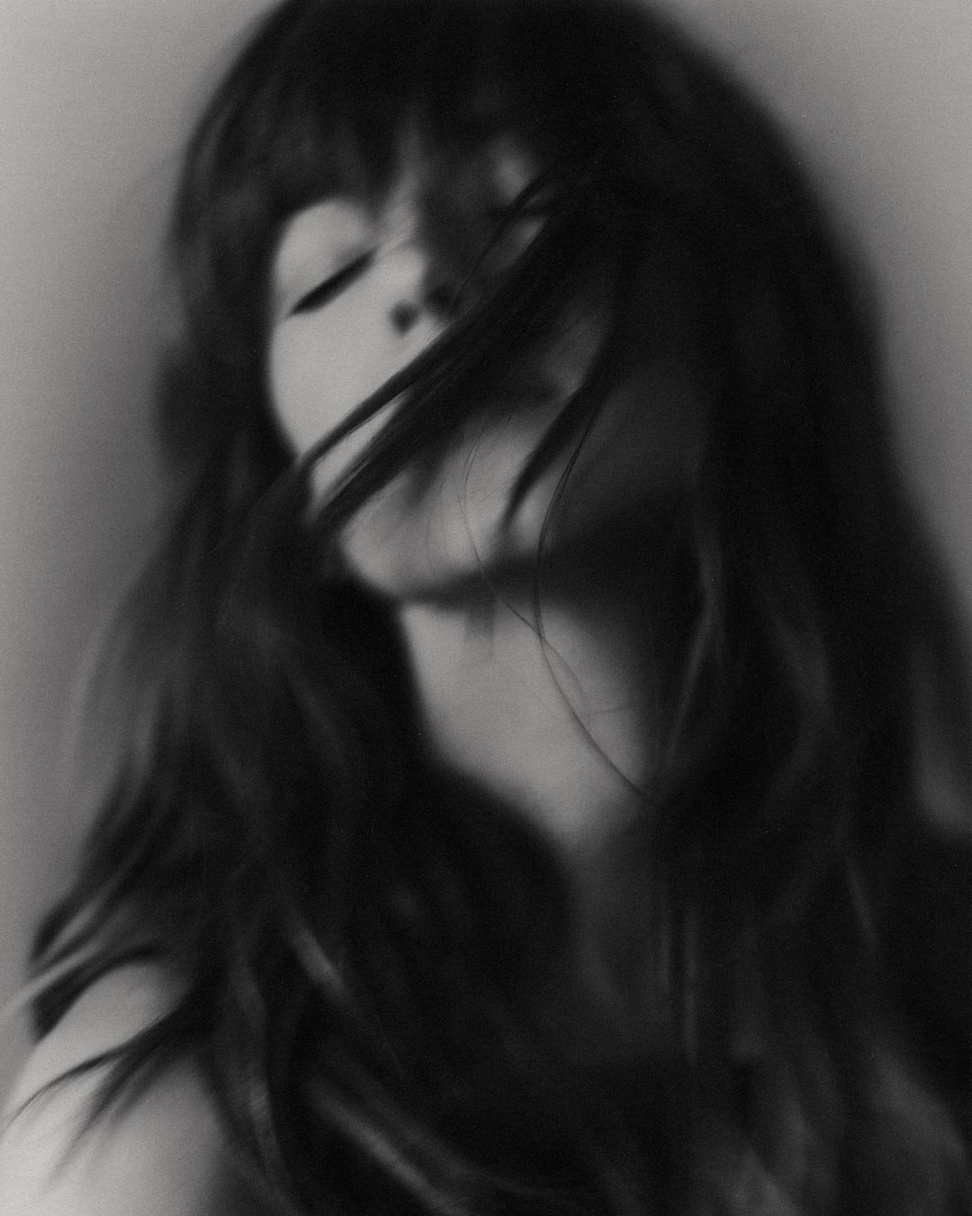Black and white portrait of a woman with long dark hair blowing across her face, eyes closed, against a plain light background.