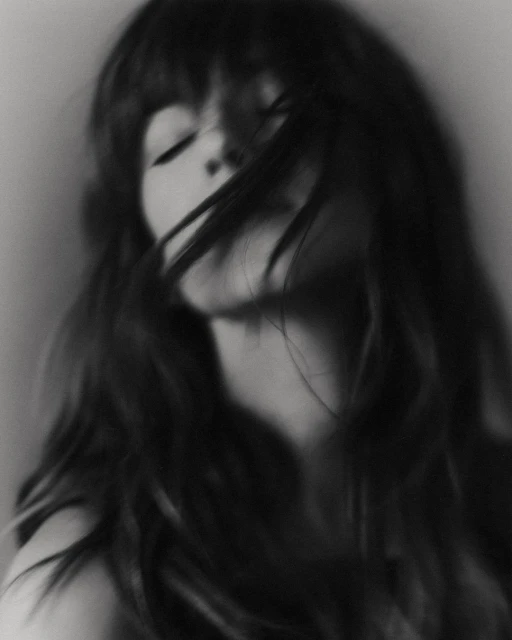 Black and white portrait of a woman with long dark hair blowing across her face, eyes closed, against a plain light background.