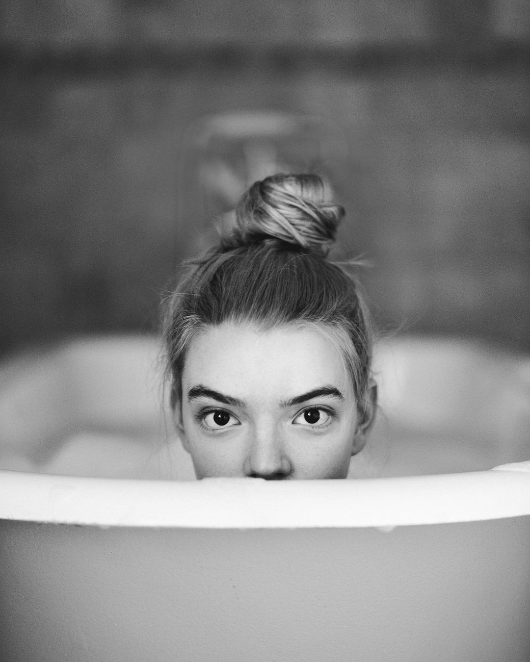 Black and white portrait of a woman in a bathtub, with focus on her face peeking over the tub's edge, gazing directly at the viewer.