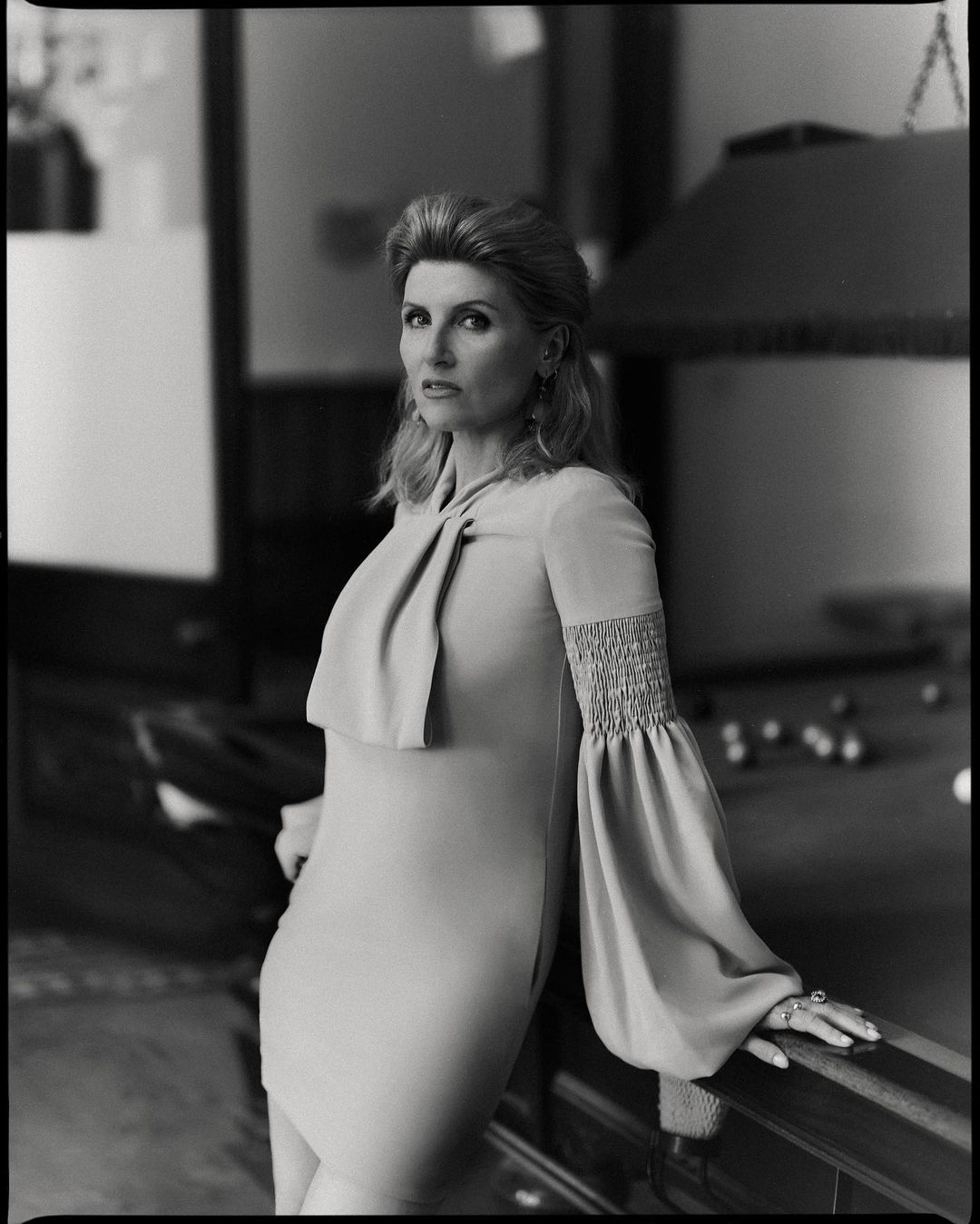 Elegant portrait of a woman in a dress with balloon sleeves leaning against a pool table in a dimly lit room.